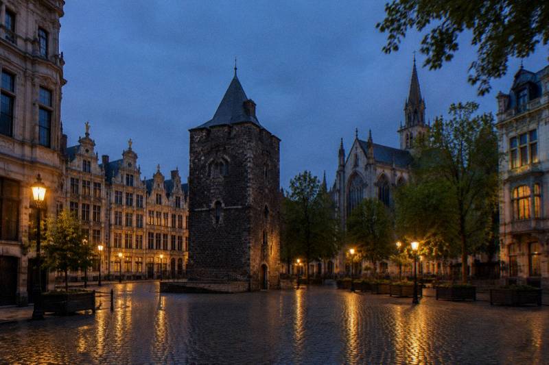 Dusk at the Black Tower on Place Sainte-Catherine in Brussels, Belgium, a short dark medieval stone tower standing at the foot of a modern church-side square, wet cobbles reflecting a few warm lamps, a deep blue sky, nobody in frame.