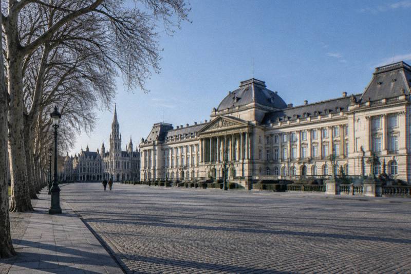 Mañana despejada en el amplio patio empedrado del Palacio Real de Bruselas, Bélgica: la larga fachada neoclásica clara bajo un cielo azul, una fila de árboles sin hojas a un lado, sombras alargadas sobre la piedra, dos figuras diminutas alejándose.
