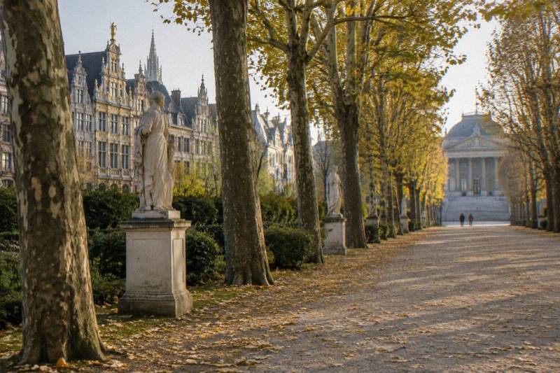 Tarde dorada de otoño en el Parc de Bruxelles en Bruselas, Bélgica: una avenida de grava bordeada de plátanos podados que dejan caer hojas amarillas, estatuas de piedra blanca sobre pedestales entre los árboles, y al fondo se vislumbra la fachada de un palacio neoclásico, con dos figuras diminutas a lo lejos.