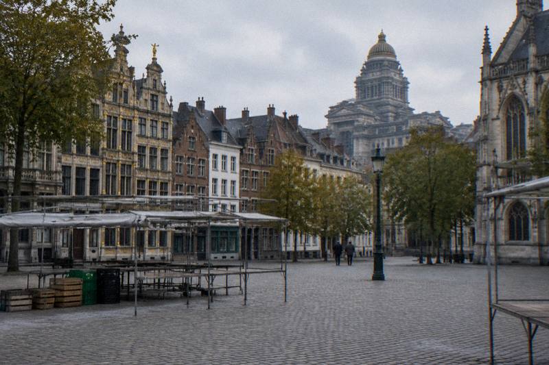 Grey weekday afternoon at the Place du Jeu de Balle in the Marolles in Brussels, Belgium, a wide cobbled square with humble brick and rendered houses around it, empty stall frames and a few crates, the huge domed Palais de Justice looming on the skyline, two tiny distant figures.