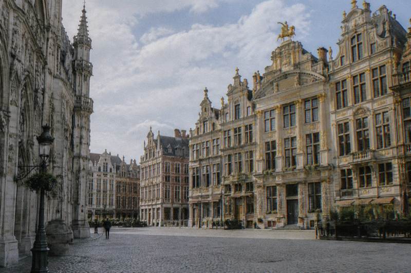 Late morning at the corner of the Grand-Place in Brussels, Belgium, ornate gilded Baroque guildhall facades with sculpted gables and gold statues leaning in over the cobbles, seen at an angle from the square's edge, a few tiny distant figures, scattered cloud.