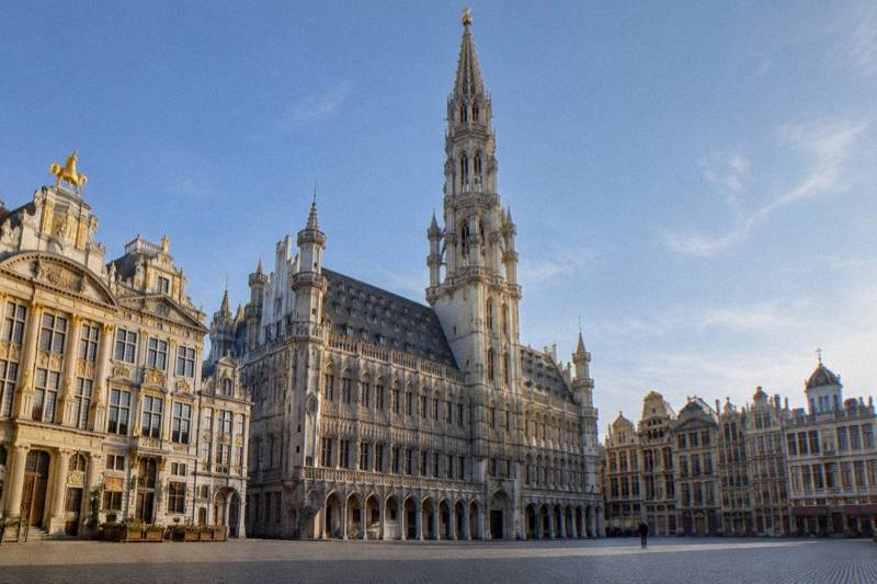 Clear morning looking straight up at the Hotel de Ville tower in Brussels, Belgium, the slender openwork Gothic spire with gold finial rising above the town hall's carved stone facade on the Grand-Place, a pale blue sky, cobbles at the bottom of the frame, one tiny distant figure.