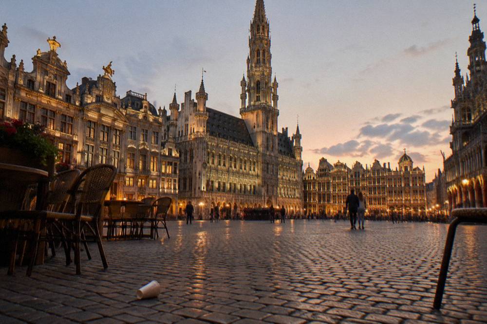 Wide city square at dusk with lit facades and a tall stone tower in the background