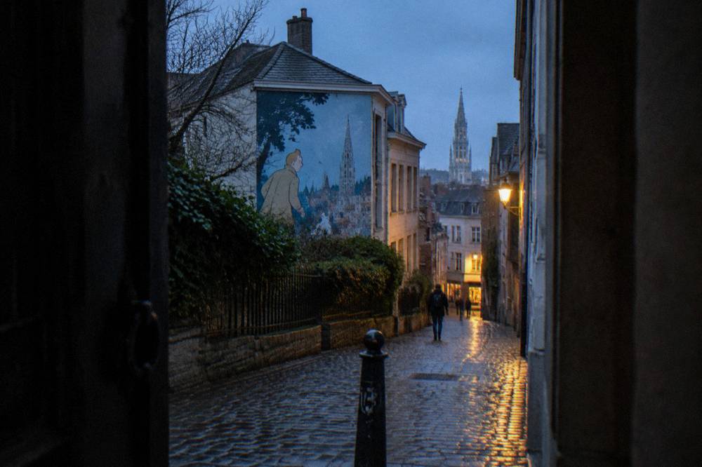 A narrow Brussels street with a painted comic mural on a building wall above the rooftops