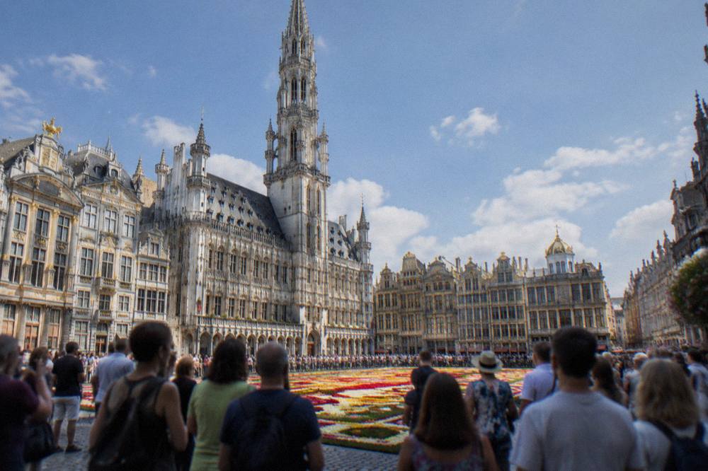 Crowds at ground level around a flower carpet pattern on the Grand-Place with the Town Hall tower above
