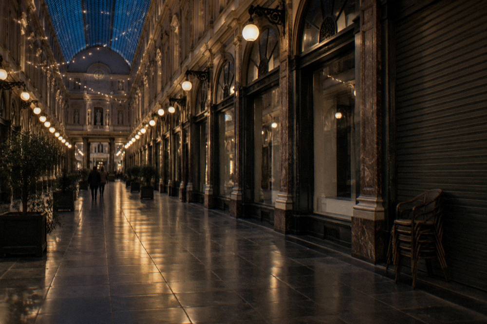 A covered glass-roofed arcade lit up at night with closed shopfronts along the gallery
