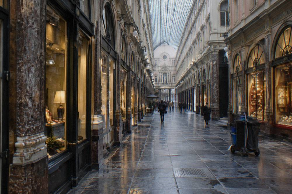 The interior of a covered shopping arcade with a arched glass ceiling and rows of illuminated shopfronts