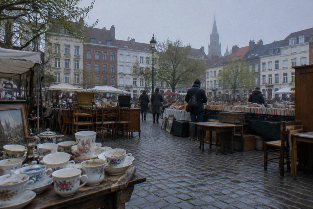 Rows of second-hand market stalls with furniture, books and household goods under open sky in a Brussels square