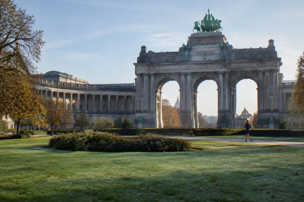 A monumental triple arch with columns and sculpture seen across a park lawn